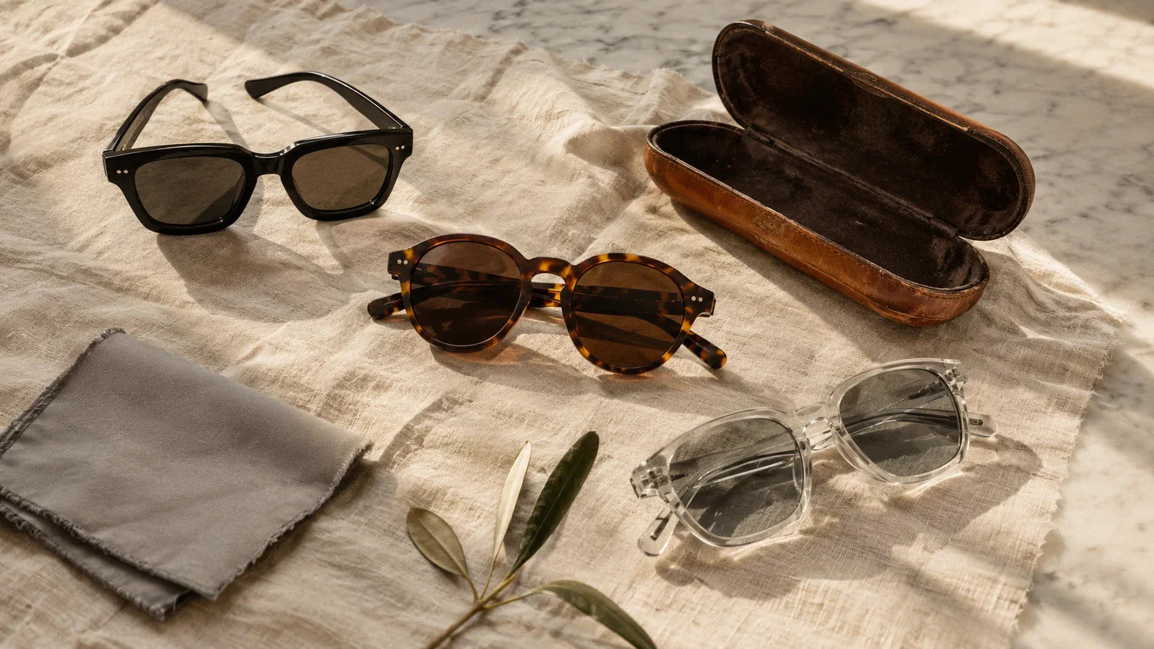 Quiet luxury sunglasses flatlay — black square, tortoiseshell round and crystal acetate frames on linen with vintage leather case on Carrara marble